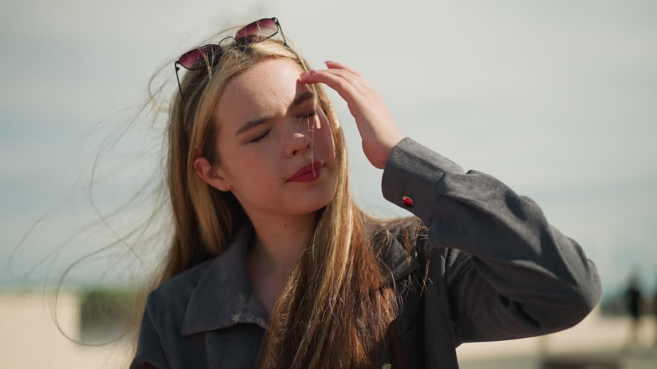 회색 옷을 입은 여성은 바람이 불면서 머리카락을 조정하고, 얼굴에 반사되는 밝은 빛으로 인해 눈이 약간 닫혀 있습니다.