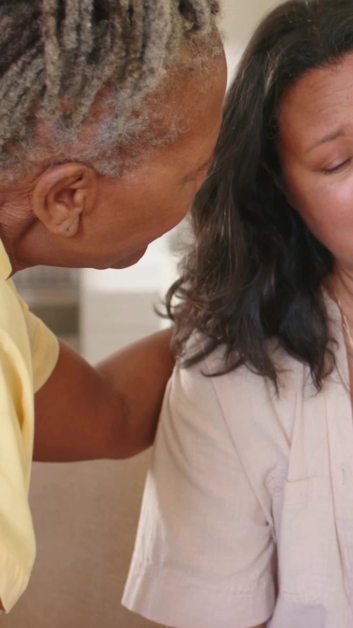Vertical video: Leaning senior whispering comfort to woman on sofa at home, friend covering hand