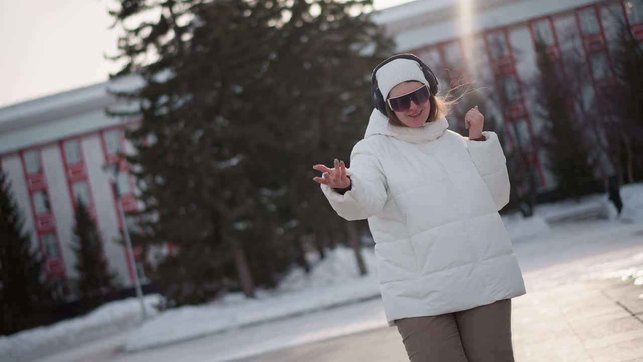 Snow dancer wearing white puffer jacket, beanie, black goggles and headphones sways hands while moving in corduroy pants and platform shoes to rhythm on snowy urban plaza under winter sunlight