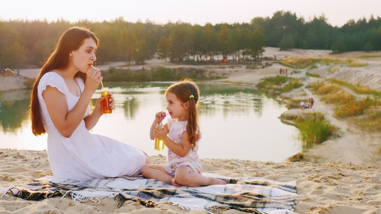Mor og datter drikker juice fra en flaske med sugerør, sidder på stranden med baggrund af