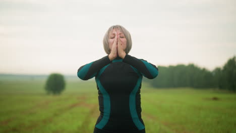 woman with arms raised brings hands together in meditation, practicing mindfulness outdoors in a grassy misty field under a cloudy sky in the background