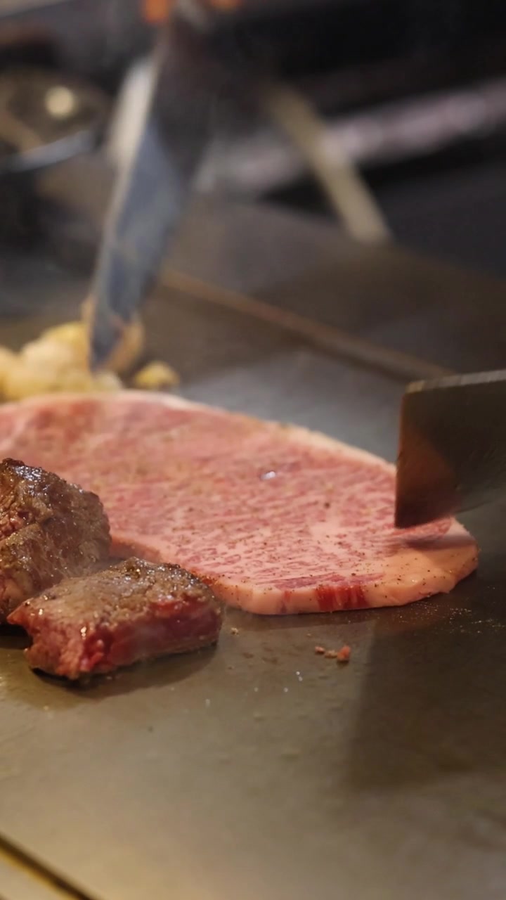 A chef uses a spatula and knife to prepare marbled steak on a sizzling grill.