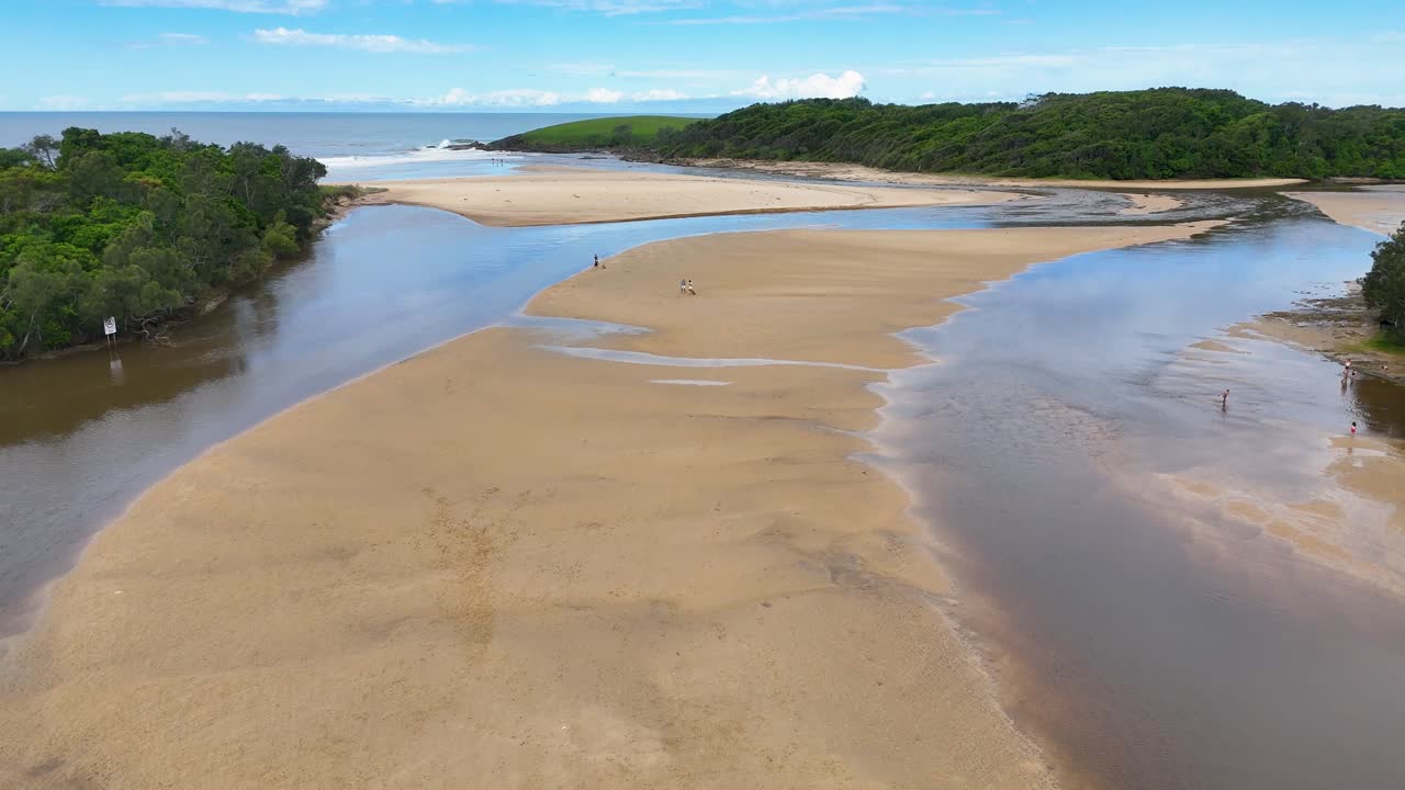Drone captures people walking on sandy estuary at Moonee Beach, bright daylight, smooth camera movement