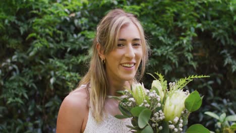 Portrait-of-happy-caucasian-woman-holding-bunch-of-flowers-in-garden-on-sunny-day