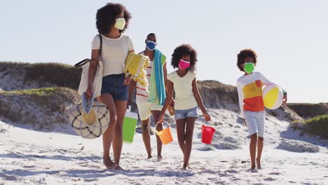 African-american-parents-and-their-children-wearing-face-masks-carrying-beach-equipment-on-the-beach