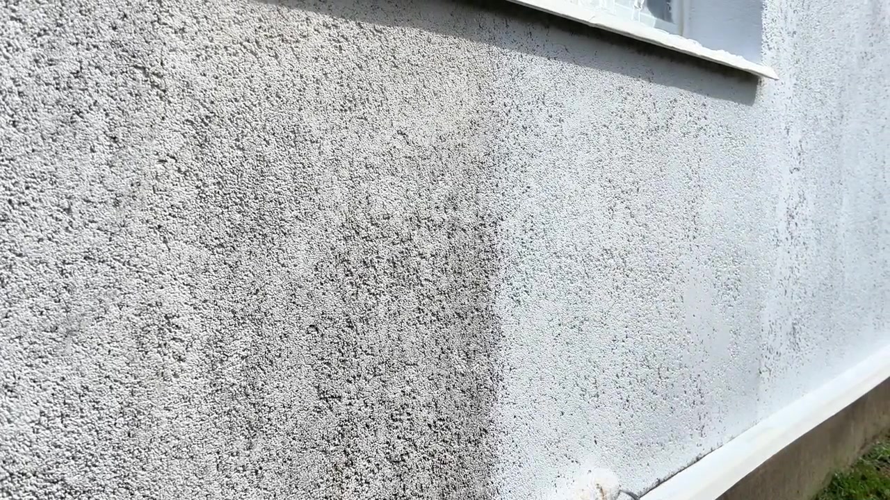 Gloved hand using a paint roller to apply white paint onto a textured exterior house wall. Ideal for home improvement, renovation, maintenance, and DIY tutorials.