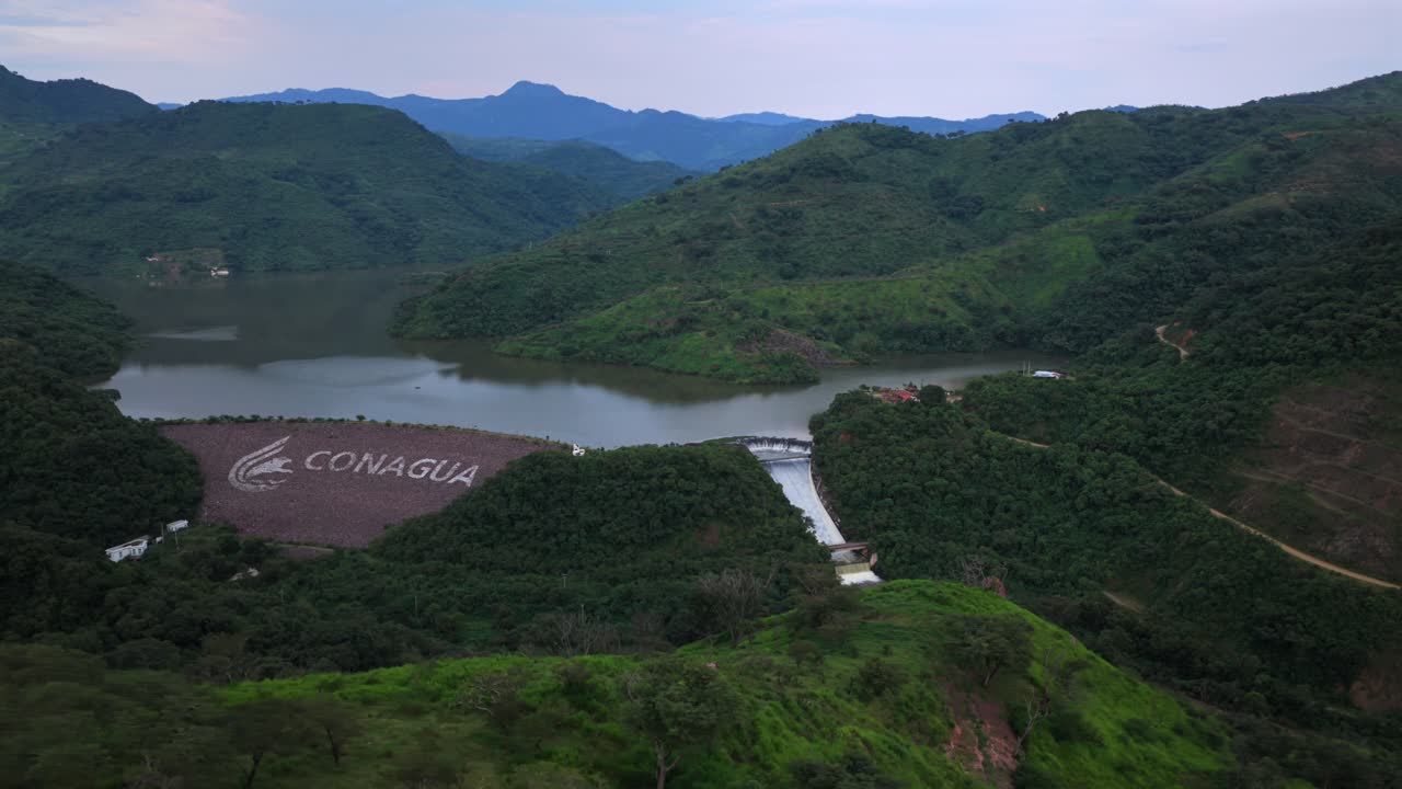 Drone video capturing the serene Presa EL Carrizo enveloped by Jalisco's lush green mountains. Overhead view highlights the tranquil dam under an expansive sky