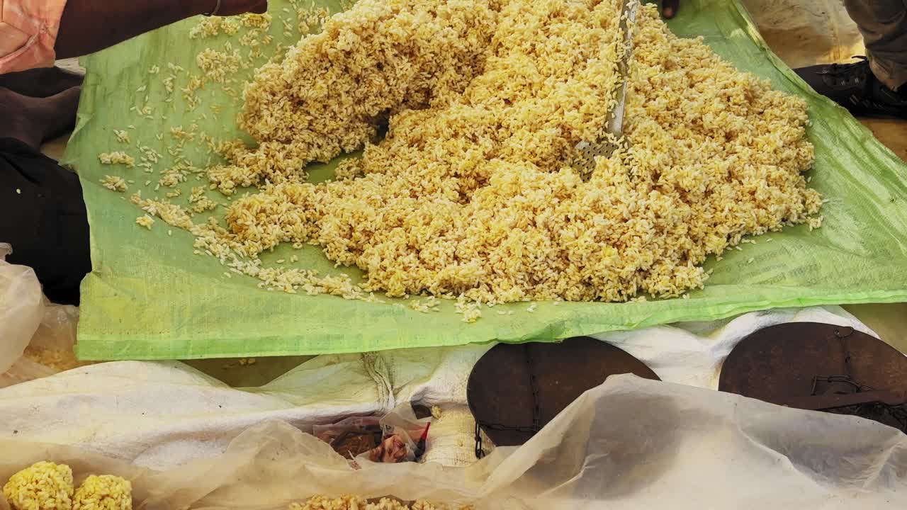 Lähikuva murmura ladoon valmistuksesta, jossa käytetään sulatettuun jaggery-siirappiin sekoitettua pörröriisiä perinteisellä alustalla, näyttäen kädet, rakenteen ja aidon intialaisen makeisen valmistuksen.