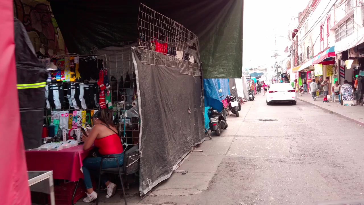 Mercado de rua movimentado mostra barracas de roupas coloridas cobertas com lonas azuis, carros estacionados, pedestres, lojas, fios elétricos e tráfego urbano ao longo de uma rua estreita na cidade do México