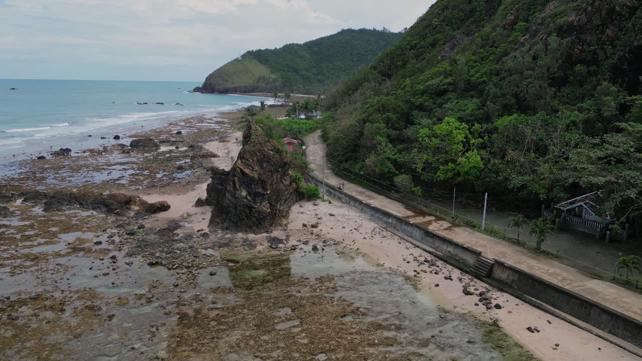 A stunning wide drone shot reveals a verdant mountain beside the shoreline, with a scenic coastal road weaving through the landscape.