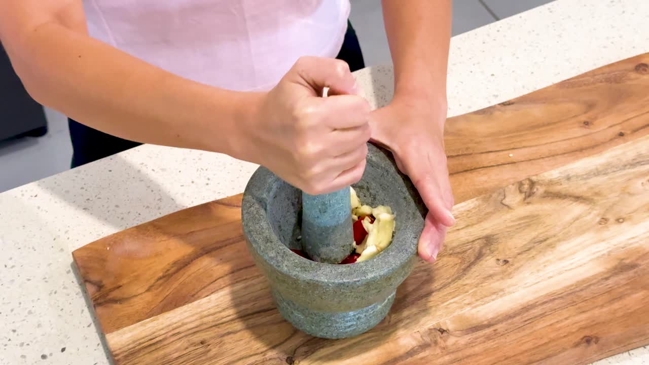 Hands vigorously grind garlic and red chili in a stone mortar and pestle on a wooden cutting board under bright, even kitchen lighting with a steady overhead camera angle