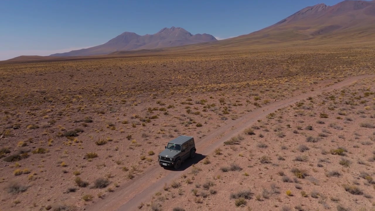 SUV compatto fuoristrada che attraversa una pista sterrata nel deserto con montagne sullo sfondo, ripreso da drone in orbita