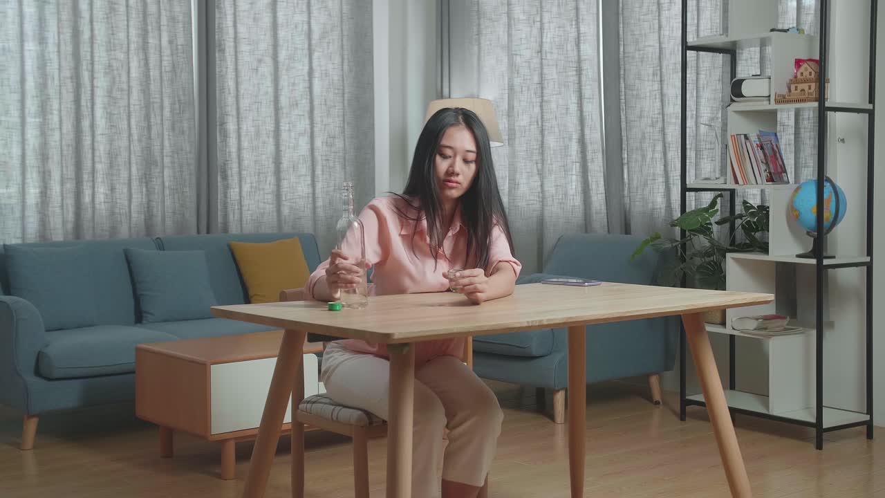 술에 취한 아시아 여성이 테이블 위에 스마트폰을 들고 보드카를 마시며 집에서 잠을 자고 있습니다.