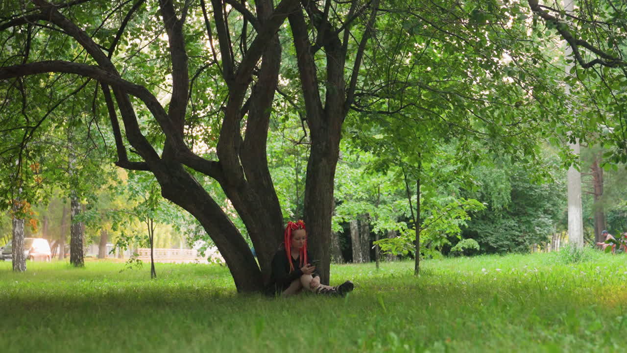 잔디밭의 큰 나무 줄기에 기대어 앉아 있는 개인, 근처에는 백팩, 편안하게 늘어진 팔다리, 얼룩덜룩한 햇빛과 시원한 그늘이 조용하고 회복적인 평온한 장면을 연출해요.