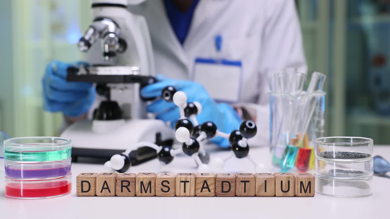 A scientist working in a laboratory with a microscope, chemical apparatus, and the word 'Darmstadtium' spelled out