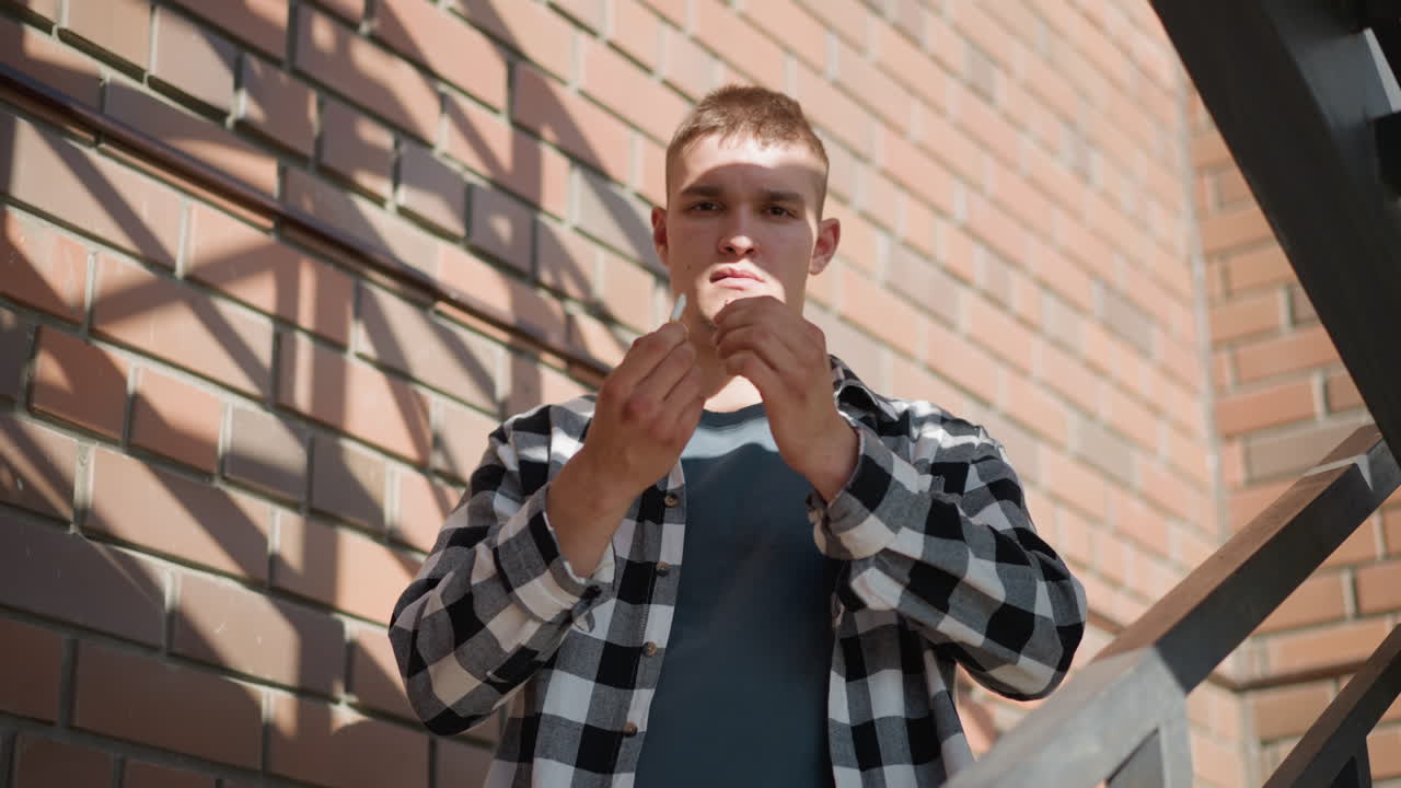 sober man in black white plaid shirt on metal staircase grips cigarette between palms bends breaks it in half with determined expression beneath sunlit brick wall backdrop