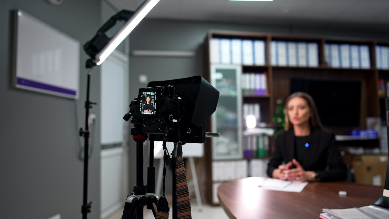 Confident serious woman wearing black jacket sits at the desk and talks. Brunette blogger records a video for her blog. Blurred backdrop.