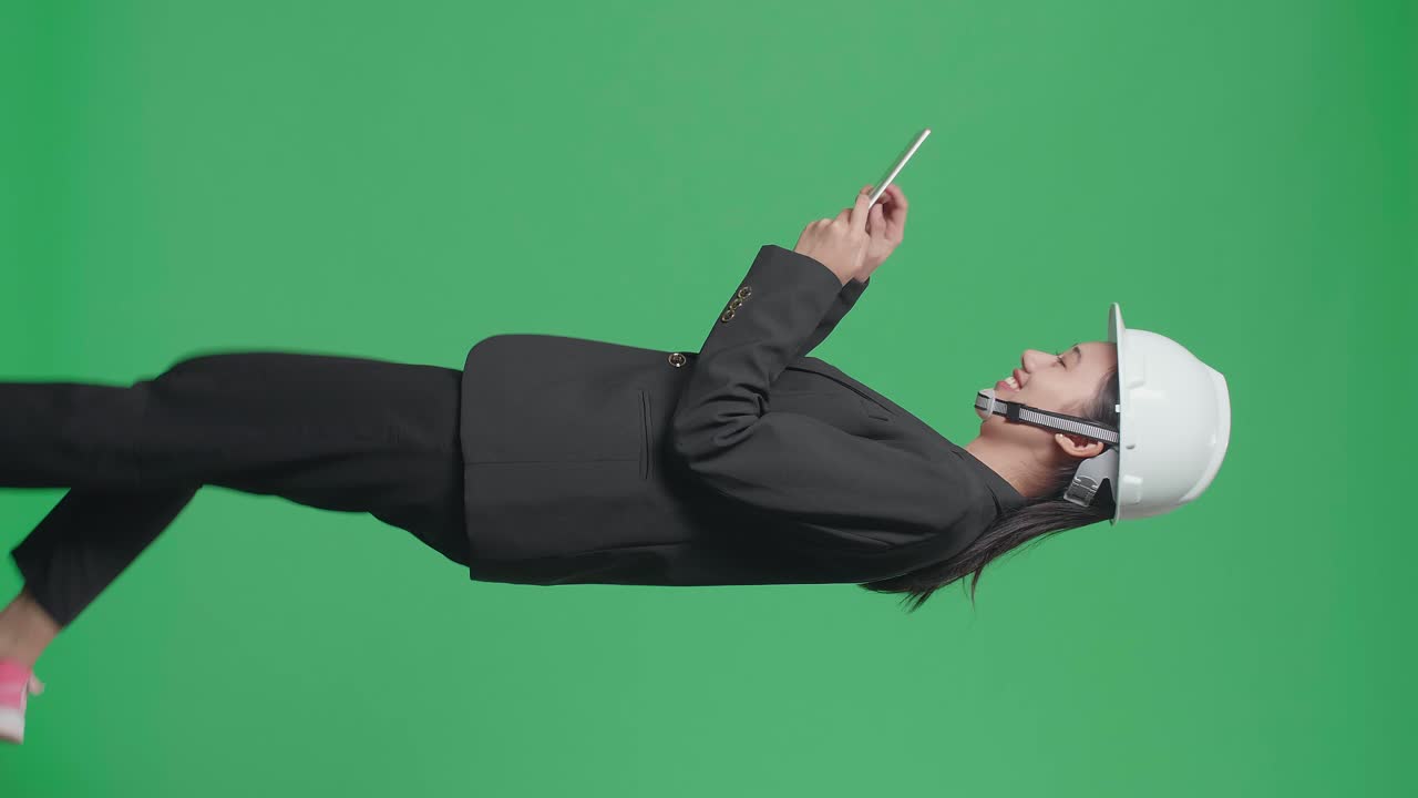 Side View Of Smiling Asian Female Engineer With Safety Helmet Using Tablet And Walking In The Green Screen Studio