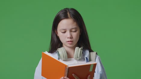 close up of unsatisfied asian teen girl student with a backpack reading book and shaking her head while standing in the green screen background studio