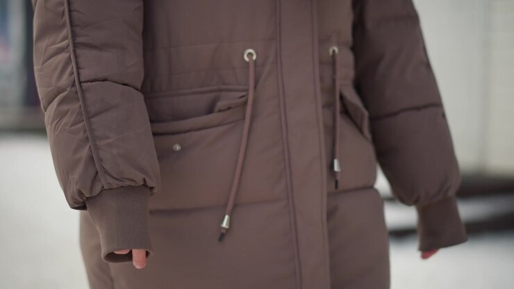 Brown winter coat focusing on details like pockets and drawstrings. fabric texture stitching seams zipper closure button fastenings are visible. Background suggests outdoor urban setting