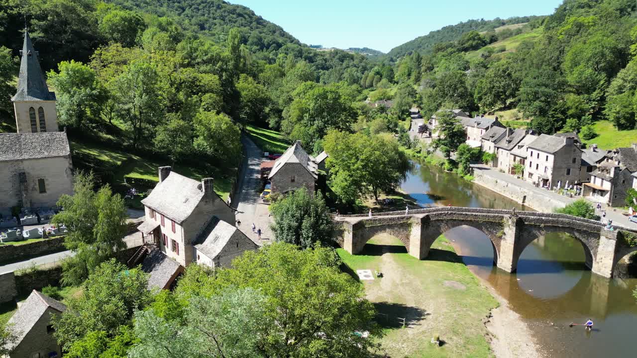 Drone aerial view in France countryside flying back from stone bridge over a river and a medieval town and church on a sunny day in Belcastel