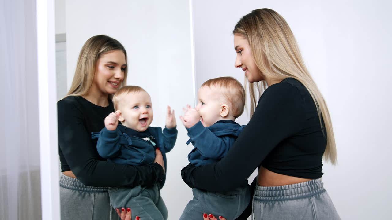 Woman with long blonde hair holding a little cute baby in front of the mirror. Happy cheerful kid touches his reflection.