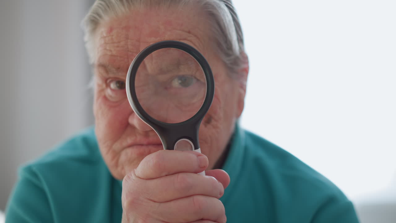 Elderly woman in green jacket holding magnifying glass, staring through lens with focused expression, sitting on bed in bright room, natural light streaming in from window, relaxed and thoughtful mood