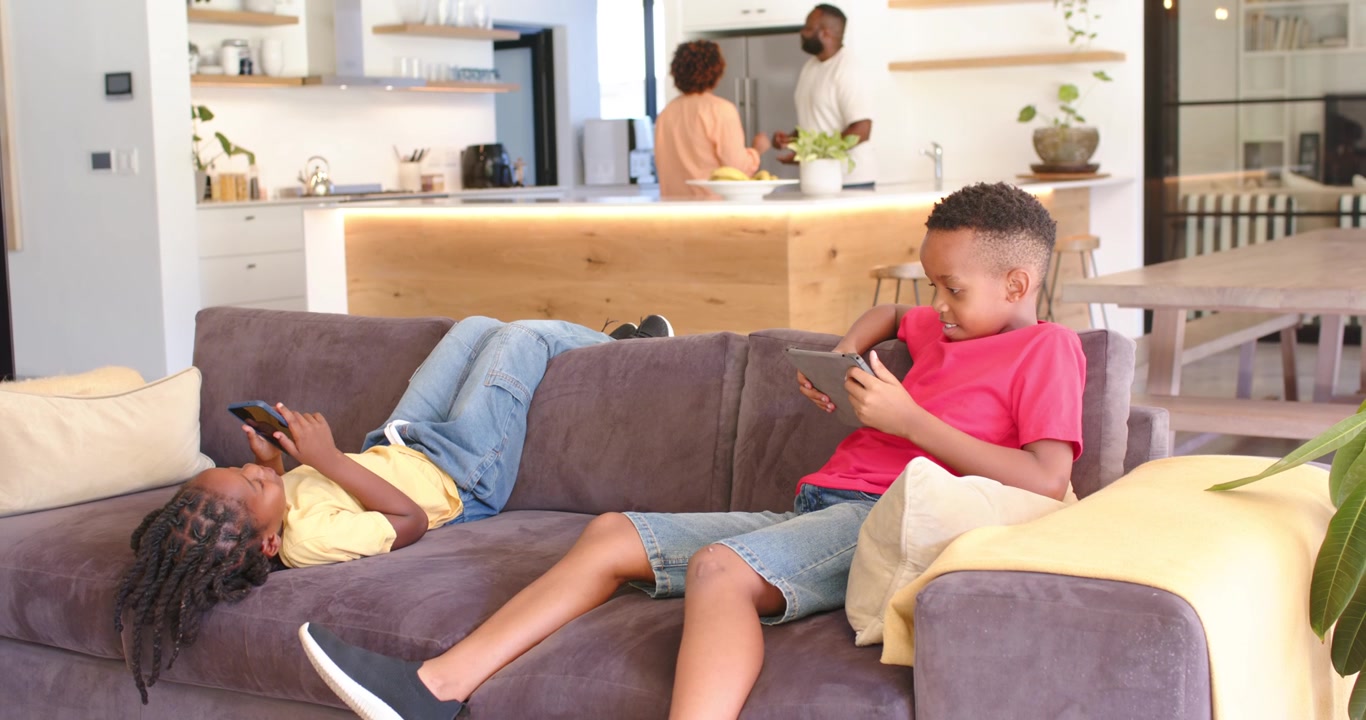Genitori afroamericani che iniziano a preparare uno spuntino e parlano mentre i bambini scorrono i dispositivi sul divano.