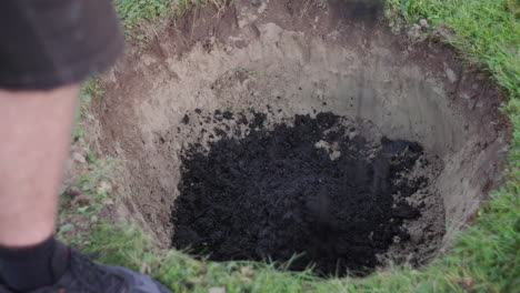 a person pours dark soil into a freshly dug hole on a lawn, with green grass surrounding the excavation.