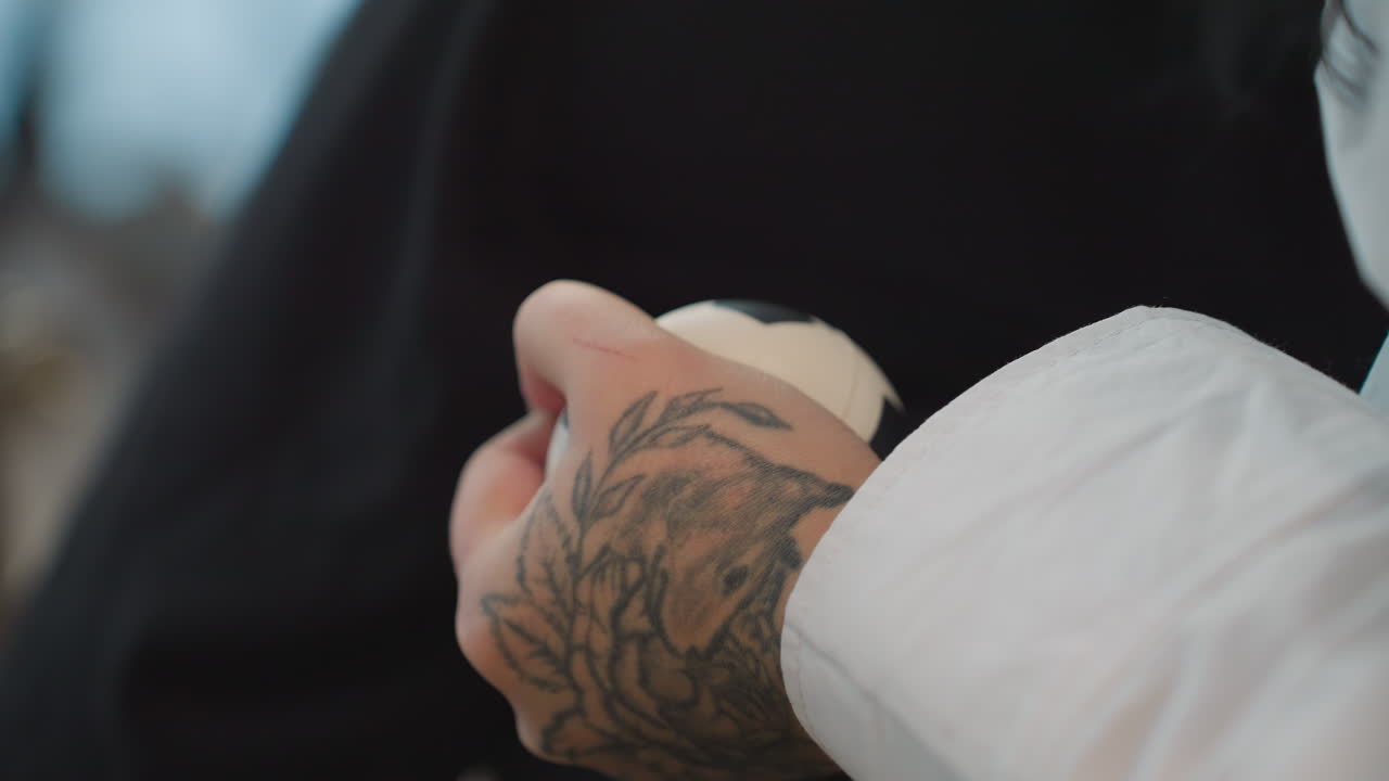 Close up of tattooed hand with visible wound squeezing stress ball firmly against black fabric, capturing emotional tension and physical strain in calm indoor setting with soft natural lighting