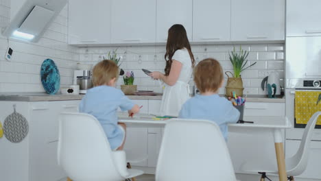 mom in the kitchen washing dishes and two sons sitting at a desk drawing on paper with colored pencils.
