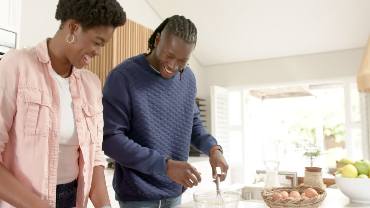 Coppia afroamericana sorridente che rompe le uova e prepara un pasto insieme in cucina a casa.