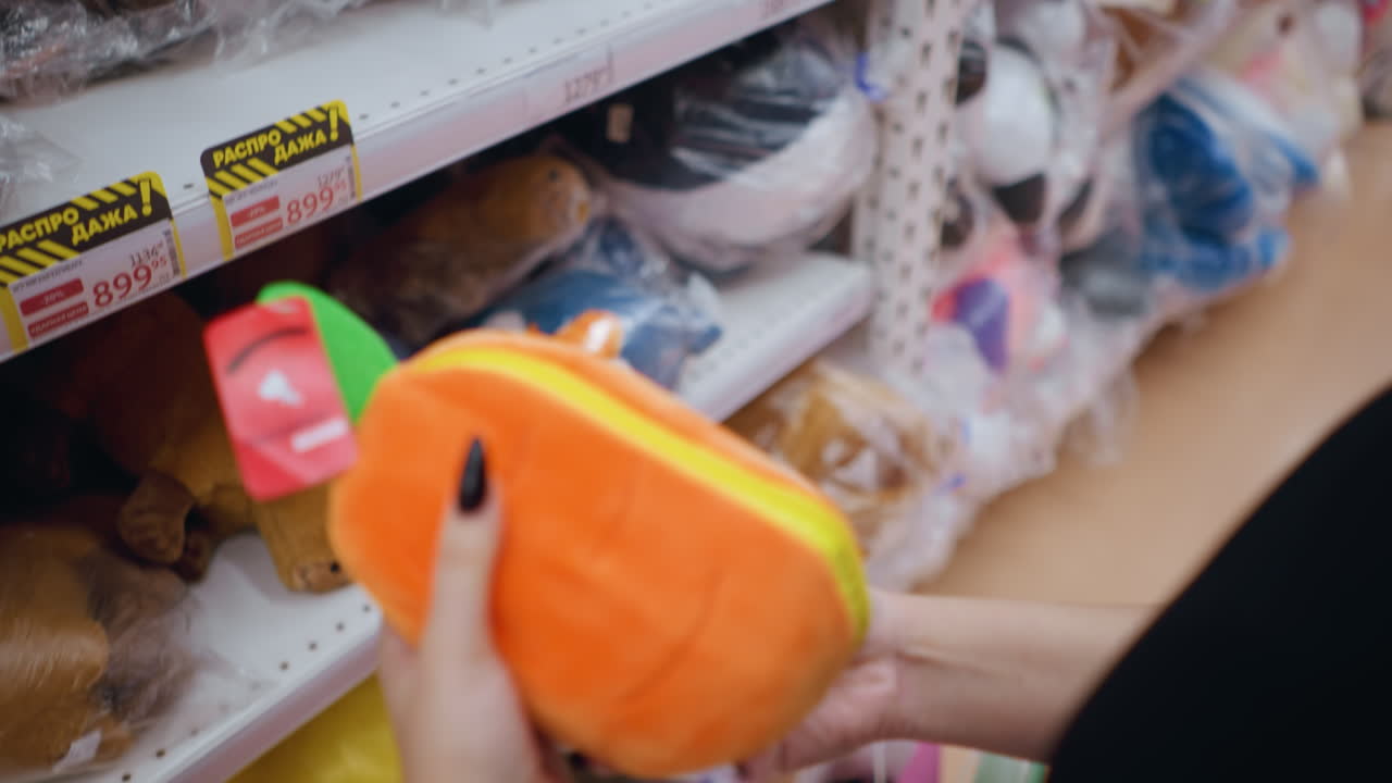 primer plano de una mujer con camiseta negra recogiendo e inspeccionando un vibrante bolso de peluche naranja con detalles verdes en un centro comercial bien iluminado, examina cuidadosamente la textura suave, los estantes de las tiendas llenos de juguetes de peluque