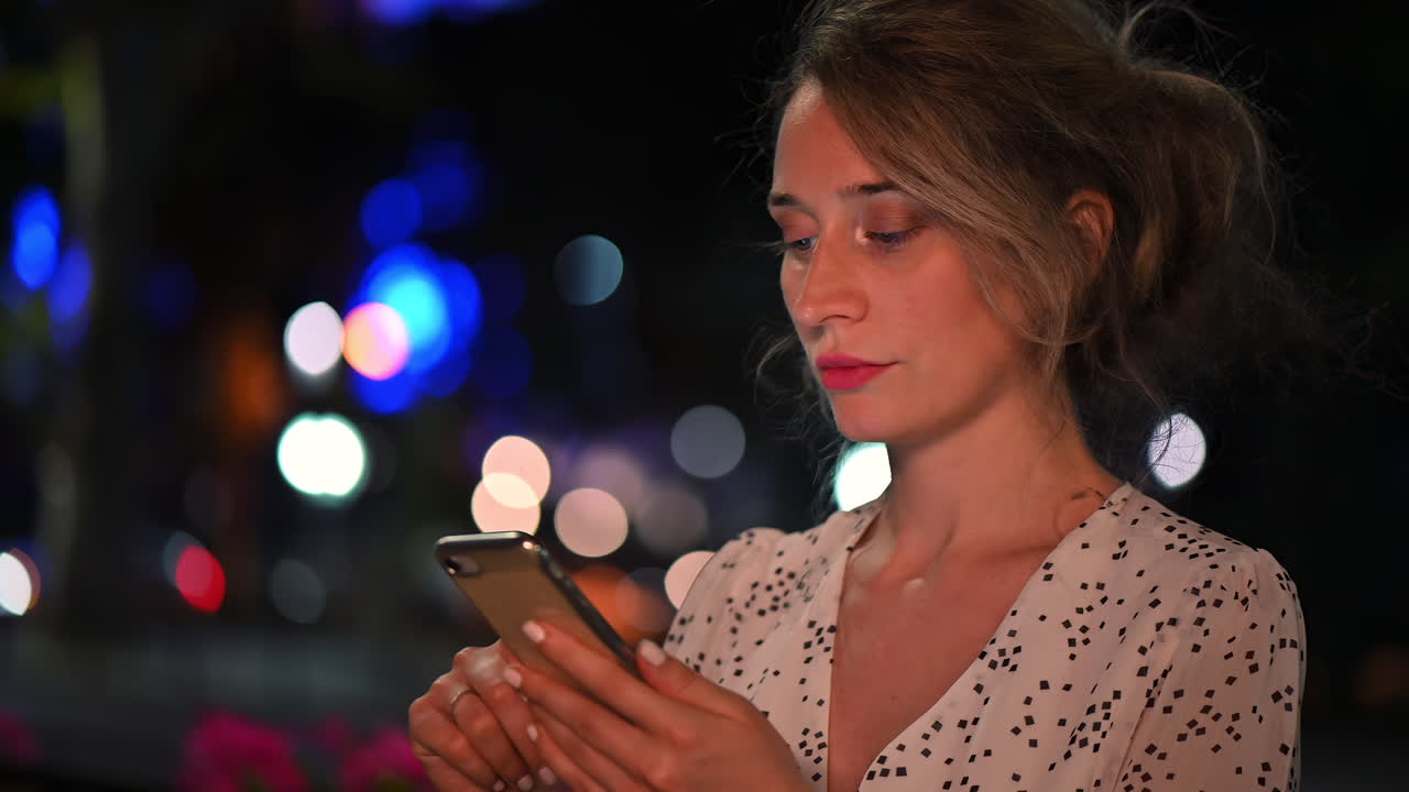 Una donna è in piedi all'aperto in una vivace città di notte, concentrata sul suo dispositivo mobile mentre luci colorate illuminano l'ambiente circostante, creando un'atmosfera vivace.