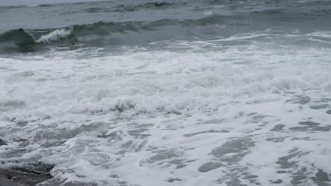 Turbulent-foamy-waves-in-slow-motion-reaching-their-end-of-life-on-the-beach-shot-from-beach-rock