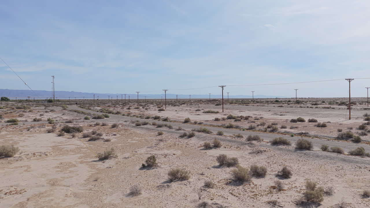 Inquadratura bassa da drone che vola in avanti sopra strade vuote a Salton City, California, verso file di linee elettriche, con montagne visibili in lontananza.