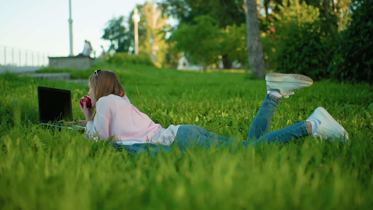 활기찬 초록색으로 둘러싸인 평온한 공원 환경에서 노트북을 사용하여 야외 잔디에 누워있는 젊은 여성, 은, 평화로운 환경에서 편안한 자세와 캐주얼 옷으로 화면에 초점을 맞추고 있습니다.
