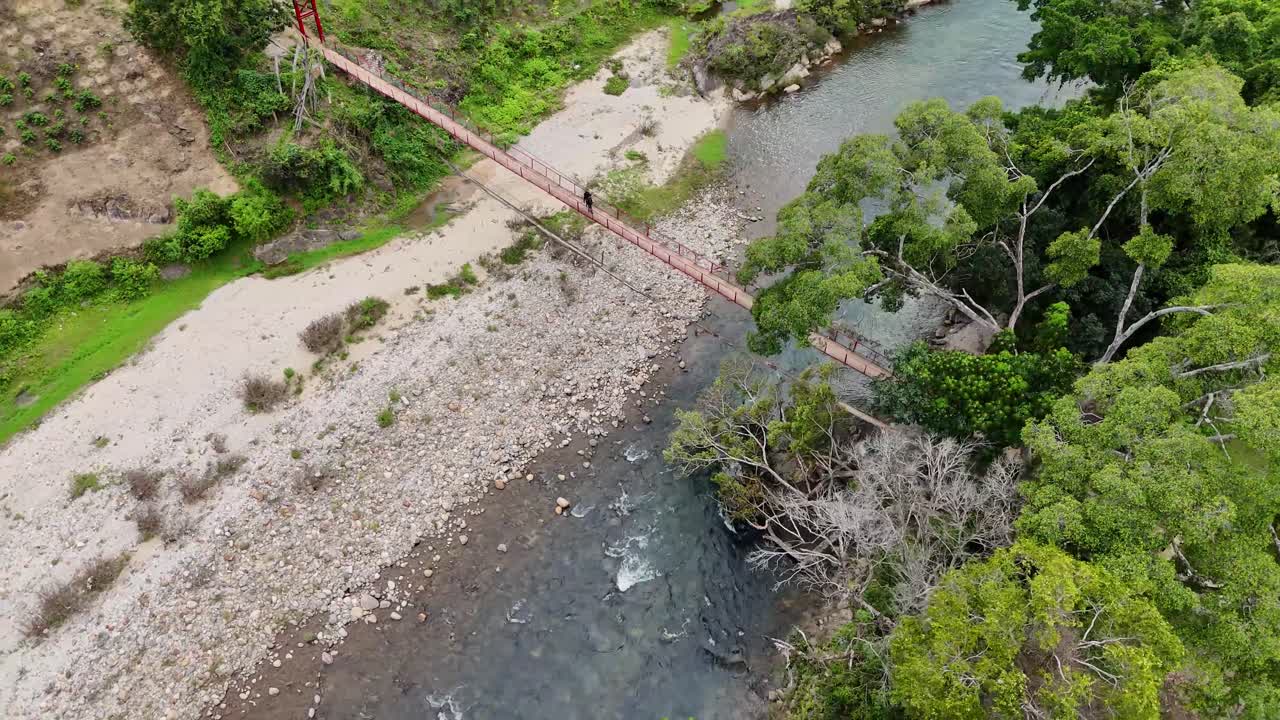 Narrow suspension bridge spans blue river with rocky bank and surrounding forest, creating a striking contrast between natural landscape and human structure captured in aerial view