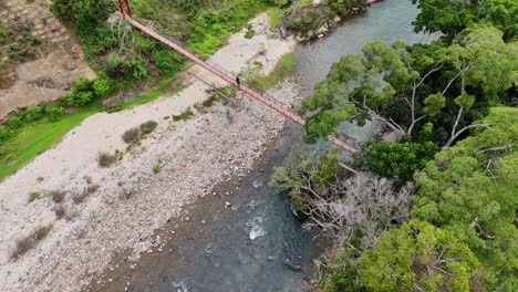 narrow suspension bridge spans blue river with rocky bank and surrounding forest, creating a striking contrast between natural landscape and human structure captured in aerial view