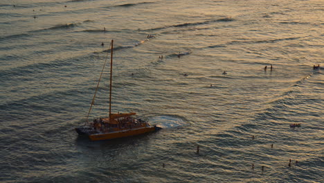 Gelber-Katamaran,-Der-Segel-Bei-Goldenem-Sonnenuntergang-Im-Seichten-Wasser-Setzt,-Mit-Touristen,-Die-Wellen-Genießen,-Hawaii