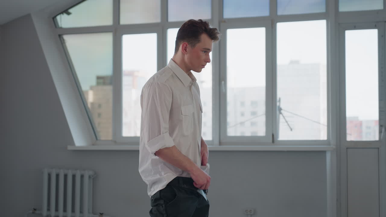 Side view showing young man gently tucking crisp white shirt into tailored trousers beside large sunlit loft window highlighting morning routine precision care and attention in modern urban apartment