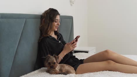 hermosa mujer joven, con pijama, se sienta en la cama con un yorkshire terrier, enviando mensajes de texto en su teléfono inteligente
