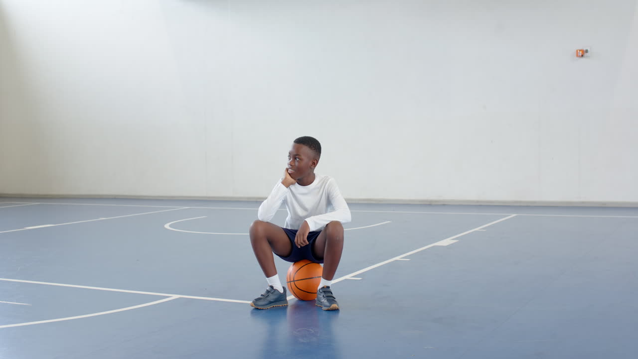 african american boy sitting on basketball in gym, looking thoughtful and relaxed, copy space
