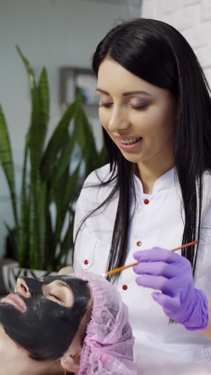 A beautiful cosmetologist applies a mask to clean the pores on the face of the client and communicates with her during the procedure at the beauty center. Vertical video