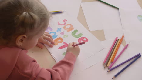 top view of blonde little girl drawing the phrase save the earth on a paper on a table in craft workshop 1