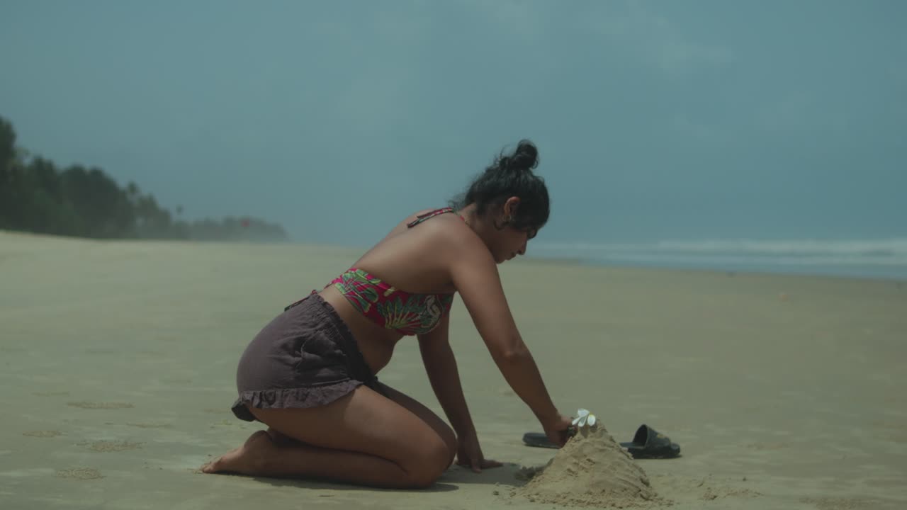 Dolly in shot of a young woman kneeling on a tropical beach, sculpting a sandcastle near the ocean waves, with sandals placed beside her in the sand, evoking a relaxed and playful summer atmosphere