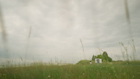a drawer, wearing a hat and checkered shirt, is intently focused on painting on a board in the middle of a lush grass field under a cloudy sky. nearby, a woman sits comfortably on a chair