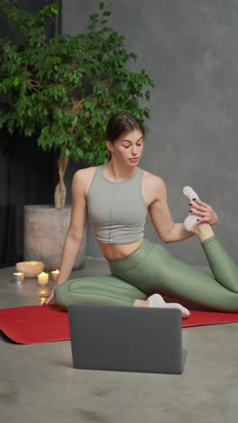 vertical video of a confident brunette girl in a sports top and green pants sitting on a red mat and doing sports and stretching with the help of online lessons through her gray laptop at home in a modern apartment near burnt candles and a large indoor plant