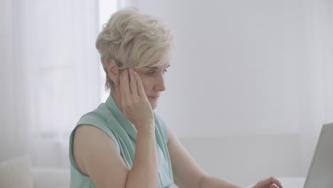 middle-aged woman with migraine is working with notebook in office typing on keyboard touching forehead by hands