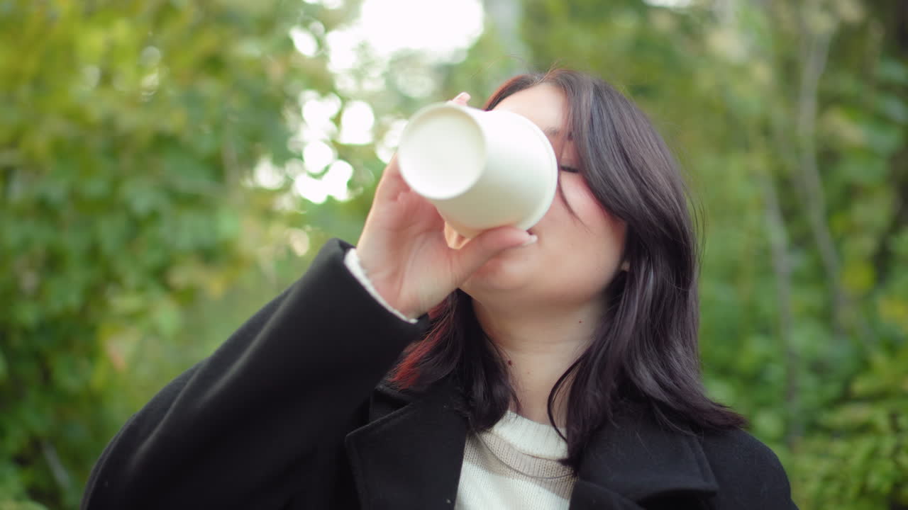 Medium close view of thoughtful young woman smiling with eyes on book, seated outdoors in autumn arboretum, black coat and striped sweater, soft greenery behind, gentle sunlight during quiet reading
